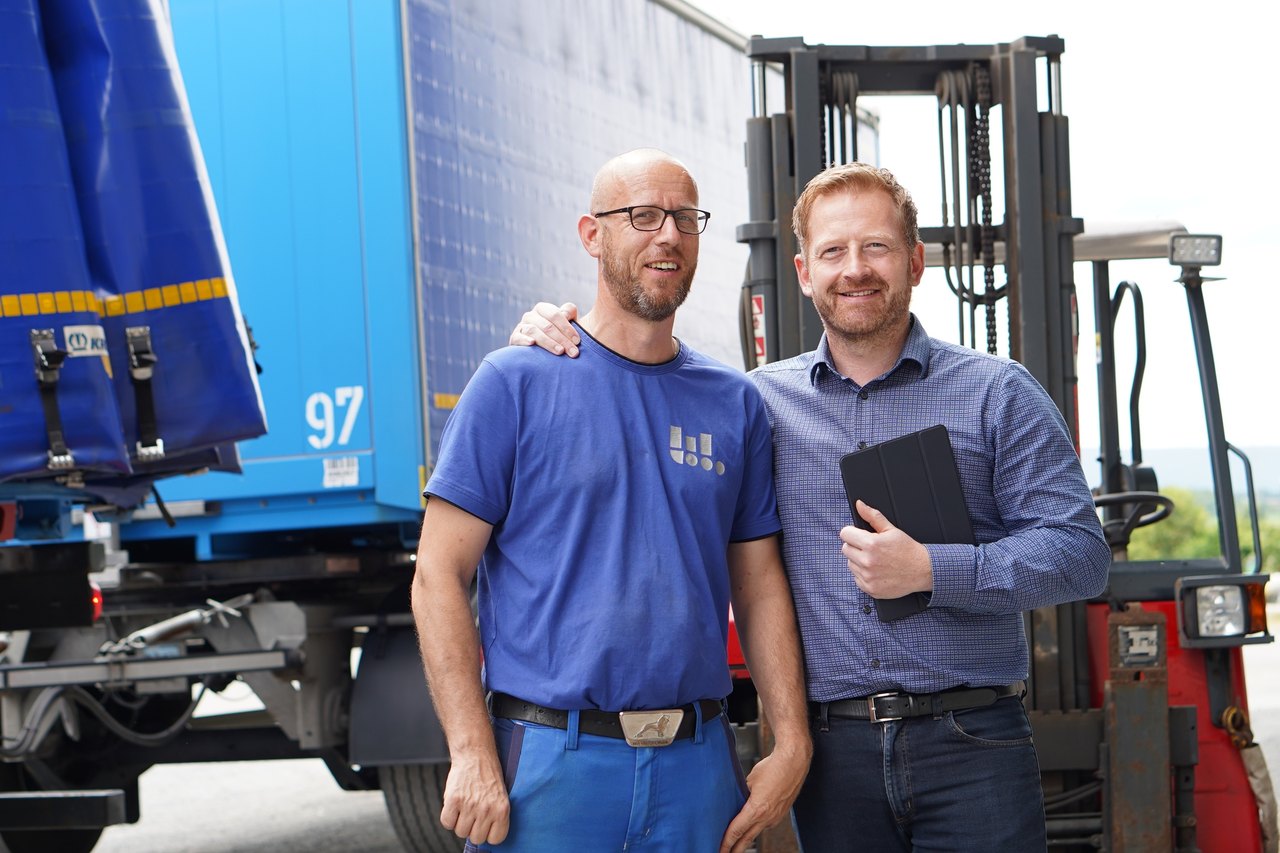 Zwei lächelnde Männer vor einem blauen Lastwagen und einem Gabelstapler.